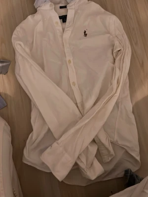 Vit skjorta från Ralph Lauren - Klassisk vit skjorta från Ralph Lauren med broderad logga på bröstet. Skjortan har lång ärm, knappar framtill och är tillverkad i mjuk bomull. Perfekt för en clean och stilren look. Har lite slitage på handen men inget som syns 