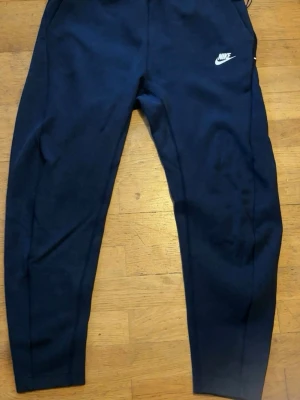 Mörkblå mjukisbyxor från Nike - Snygga mörkblå mjukisbyxor från Nike med klassisk vit logga på låret. Byxorna har en smal passform och är tillverkade i mjukt och bekvämt material, perfekta för chill eller träning. Elastisk midja för skön passform.