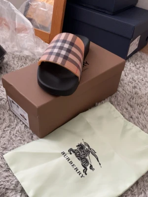Burberry rutiga slides i svart - Säljer ett par snygga slides från Burberry med klassiskt beige, svart och rött rutmönster över foten. Sulan är svart och har Burberry-loggan präglad. Perfekta för en trendig och avslappnad look. Kommer med originalpåse och låda.