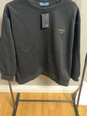 Svart sweatshirt från Prada - Säljer en svart sweatshirt från Prada med klassisk rund halsringning och ribbade muddar. Tröjan har en ikonisk metalltriangel med Prada-logga på bröstet. Tillverkad i mjukt bomullsmaterial som känns riktigt skönt mot huden. Perfekt för dig som gillar stilrena designer. En stor S 
