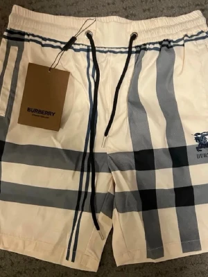 Rutig Burberry shorts med snörning - Säljer ett par klassiska Burberry shorts i beige med svart och grått rutmönster. Shortsen har elastisk midja med svart snörning och sidofickor. Perfekta för sommaren och har den ikoniska Burberry-loggan broderad på ena benet.