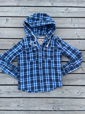 Blårutig hollister overshirt! - Snygg blårutig skjorta från Hollister med huva och dragsko. Skjortan har två bröstfickor med lock och knappar framtill. Den är skadefri, fråga vid intresse!! Den är S men sitter mer som en XS!