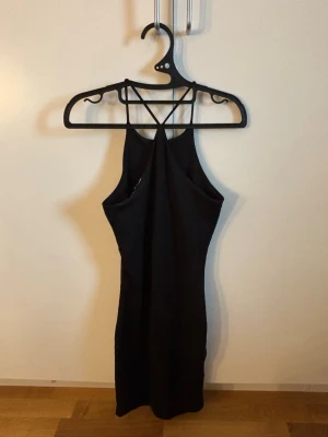 Svart tight knälång klänning - Säljer en svart tight knälång klänning med smala axelband och snyggt korsad rygg. Klänningen har en minimalistisk design och är perfekt för dig som gillar en enkel men stilren look. Materialet känns mjukt och stretchigt.