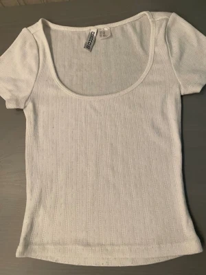 Vit ribbad topp från H&M - Säljer en vit ribbad topp från Cropp med korta ärmar och djup rundad urringning. Toppen har ett diskret hålmönster i tyget och är tillverkad i ett mjukt, stretchigt material som sitter snyggt på kroppen. Perfekt till jeans eller kjol.