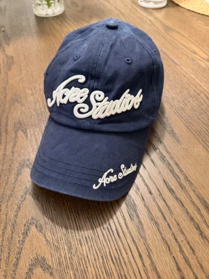 Mörkblå keps från Acne Studios - Snygg mörkblå keps från Acne Studios med vit broderad logga både framtill och på skärmen. Klassisk form med justerbar rem baktill och ventilationshål. Perfekt för att lyfta din streetstyle-look.