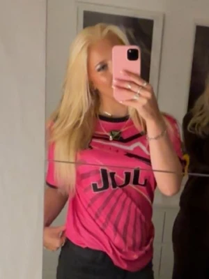 Rosa grafisk t-shirt från JUL - Säljer en rosa t-shirt från JUL med svart och gul grafiskt tryck framtill. T-shirten har korta ärmar och en rund halsringning. Perfekt för dig som vill sticka ut med en färgstark och unik stil.
