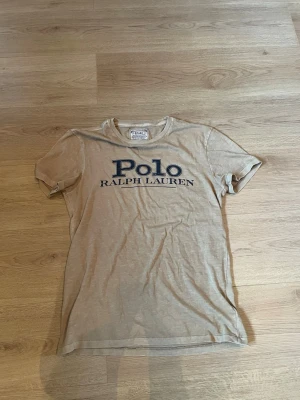 Beige t-shirt från Polo Ralph Lauren - Snygg beige t-shirt från Polo Ralph Lauren med stor blå logga på bröstet. Modellen är custom slim fit och har korta ärmar. Tillverkad i mjuk bomull som känns skön mot huden. Perfekt för en avslappnad och stilren look.                                       Pris kan diskuteras vid snabb affär 