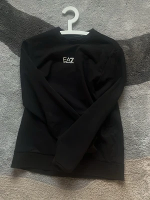 Svart sweatshirt från EA7 Emporio Armani - Svart sweatshirt med rund hals från EA7 Emporio Armani. Tröjan har en stilren design med vit logotyp på bröstet och ribbade muddar vid ärmslut och nederkant. Perfekt för en avslappnad och trendig look.