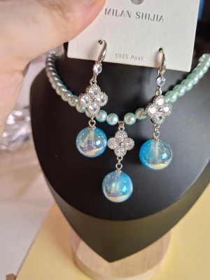 Blått örhängen halsband  - Snyggt smyckeset med halsband och örhängen från Milan Shijia. Setet har ljusblå pärlor och gnistrande klara stenar i blommönster. Hängsmyckena är klotformade i blått och vitt med skimrande effekt. Perfekt för dig som gillar unika accessoarer.
