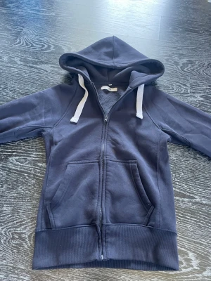 Mörkblå hoodie med dragkedja från Russemerch - Mysig mörkblå hoodie med vit snörning i huvan och dragkedja framtill. Hoodien har två stora fickor och är fodrad med mjuk fleece på insidan. Perfekt för chill dagar eller när du vill vara extra bekväm.