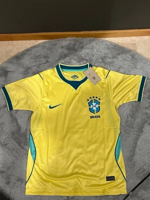 Brasilien Vm 2026 fotbollströja  - Gul Brasilien fotbollströja från Nike med blå detaljer och CBF-emblem på bröstet. Tröjan har korta ärmar, rund hals med blå kant och är tillverkad i ett lätt, ventilerande material perfekt för träning eller match.