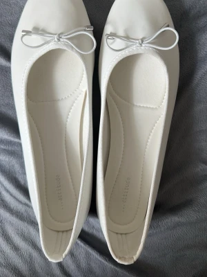 Vita ballerinas med rosett från Attitude - Säljer ett par vita ballerina skor från Attitude med rund tå och en söt rosett framtill. Skorna har en klassisk och stilren design, perfekt för dig som gillar enkelhet och elegans. Materialet är syntet och de är lätta att matcha med olika outfits.