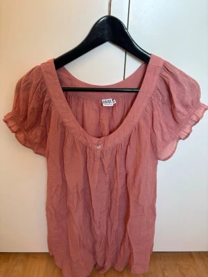 Rosa blus  - Söt rosa blus från Saint Tropez med puffiga korta ärmar och djup v-ringning. Blusen har en knappdetalj framtill och är gjord i ett lätt, luftigt material som passar perfekt till varmare dagar.