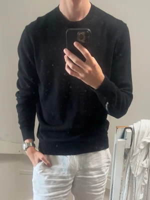 Svart rundhalsad tröja - Klassisk svart tröja med rund halsringning och ribbade muddar vid ärmslut och nederkant. Tröjan har en clean och stilren look som passar till det mesta. Perfekt för dig som gillar enkel och tidlös stil.