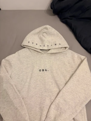 Ljusgrå hoodie från Jack & Jones - Ljusgrå hoodie från Jack & Jones med trycket 'ORG.' på bröstet och 'ORIGINALS' på huvan. Klassisk passform med känguruficka och mjukt material, perfekt för chill dagar. Snygg och enkel design som funkar till det mesta.