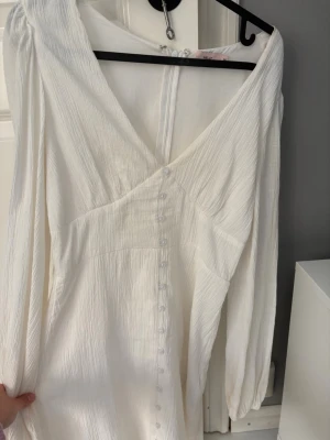 Vit långärmad omlottklänning - Vit klänning med lång ärm och v-ringning, omlott-design och knappar framtill. Tillverkad i ett lätt, krinklat tyg som ger en avslappnad känsla. Perfekt för sommarens alla tillfällen.