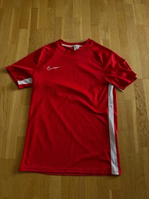 Röd Nike sporttröja med vita detaljer - Röd sporttröja från Nike med vita ränder längs sidorna och vit Nike-logga på bröstet. Kortärmad modell i funktionsmaterial, perfekt för träning eller fotboll. Snygg och sportig design 