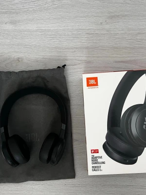 JBL 670NC - Snygga svarta over-ear hörlurar från JBL med adaptiv brusreducering och mikrofon för samtal. Kommer med en praktisk tygpåse och originalkartong. Perfekta för musik, gaming eller videosamtal. Trådlös design och bekväm passform.
