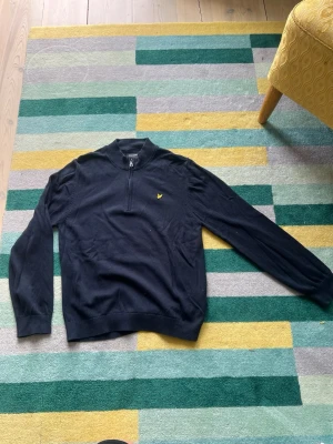 Svart halvzip från Lyle & Scott - Snygg svart tröja från Lyle & Scott med halv dragkedja och gul logga på bröstet. Tröjan har ribbade muddar och hög krage, perfekt för lager på lager. Passar dig som gillar stilren och enkel design.