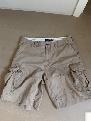 Beige shorts från Polo Ralph Lauren - Säljer ett par beige cargo shorts från Polo Ralph Lauren med flera praktiska fickor och klassisk knappstängning. Shortsens material är bomull och de har en avslappnad passform. Extremt snygga nu till sommaren där de är perfekta. Shortsen är precis som nya och har används ett fåtal gånger. Denna modell på shorts är väldigt populär och har stor efterfrågan. Storlek 36 men passar mig som är 184! Pris kan diskuteras vid snabb affär!