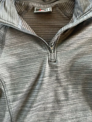 Grå träningströja från High Mountain - Snygg grå långärmad träningströja från High Mountain med halv dragkedja framtill. Tillverkad i ett mjukt och stretchigt funktionsmaterial med diskret mönster. Perfekt för träning eller utomhusaktiviteter.