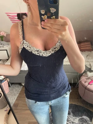 Mörkblå linnetopp från intimissimi - Snygg mlrkblå linnetopp med vit spets upptill och v-ringad hals. Toppen är ärmlös och har en elegant, feminin look som passar perfekt till jeans eller kjol. Materialet känns mjukt och följsamt mot huden.