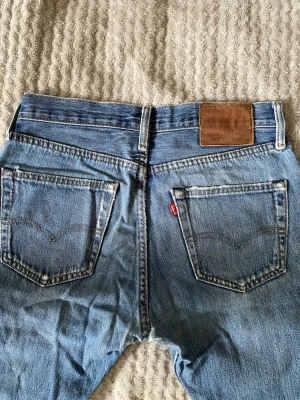 Levi's 501 ljusblå jeans vintage - Klassiska Levi's 501 jeans i ljusblå tvätt med raka ben och femficksmodell. Jeansen har den ikoniska röda Levi's-lappen på bakfickan och läderpatch i midjan. Tillverkade i slitstark bomull, perfekt för en avslappnad och tidlös look.