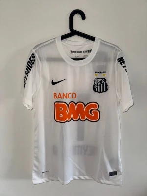 Vit Santos FC Neymar Jr matchtröja - Säljer en vit Santos FC matchtröja från Nike med Neymar Jr och nummer 11 på ryggen. Tröjan har tryckta sponsorer som BMG och Netshoes, klubbmärke på bröstet och är tillverkad i lätt, ventilerande material med Dri-FIT-teknologi. Perfekt för dig som älskar brasiliansk fotboll.