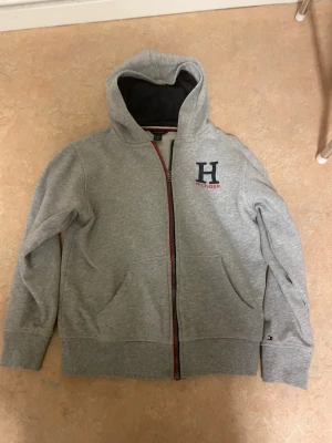 Grå hoodie från Tommy Hilfiger - Snygg grå hoodie med dragkedja från Tommy Hilfiger. Stor H-logga och Hilfiger-text på bröstet, samt liten flagga på ärmen. Mjuk bomullsblandning, två fickor fram och huva med svart innerfoder. Perfekt för en avslappnad och stilren look.
