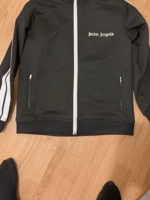 Palm Angels svart track jacket - Palm Angels svart track jacket med vita detaljer och dragkedja framtill. Jackan har vita ränder längs ärmarna, två fickor med dragkedja och logga på bröstet. Tillverkad i polyester och bomull för en sportig och clean look.Storlek:s/m