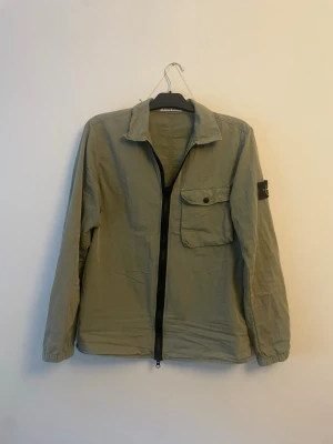 Grön overshirt från Stone Island - Snygg ljusgrön overshirt från Stone Island med klassisk patch på ärmen. Jackan har en bröstficka med knapp, dragkedja framtill och avslappnad passform. Tillverkad i lätt material som passar perfekt till lager-på-lager-stil.
