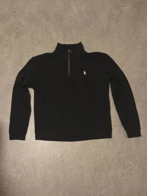 Svart half-zip tröja från Polo Ralph Lauren - Svart långärmad tröja från Polo Ralph Lauren med half-zip och hög krage. Tröjan har det klassiska vita Polo-logotrycket på bröstet och är tillverkad i mjukt material. Perfekt för en clean och stilren look.