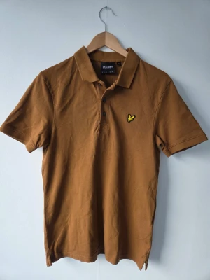 Brun pikétröja från Lyle & Scott - Snygg brun pikétröja från Lyle & Scott med klassisk krage och knappslå framtill. Tröjan har korta ärmar och den ikoniska gula loggan broderad på bröstet. Perfekt för en stilren och avslappnad look.