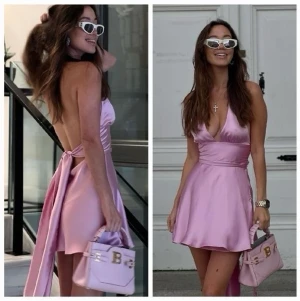 Rosa omlottklänning i satin - Snygg rosa kortklänning i satin med omlott och djup v-ringning. Klänningen har öppen rygg och knytdetalj bak, vilket ger en elegant och trendig look. Perfekt för dig som vill sticka ut med en feminin och modern stil.