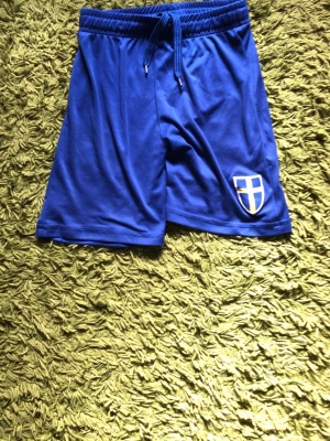Blå sportshorts med emblem - Säljer ett par blå sportshorts med resår och dragsko i midjan. Shortsen har ett vitt och gult emblem på vänster ben och är tillverkade i ett lätt och snabbtorkande material, perfekt för träning eller match.