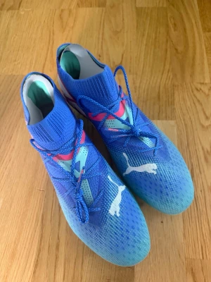 Puma Future - Säljer ett par snygga blåa Puma Future. Skorna har turkosa och rosa detaljer samt vit Puma-logga på sidan och ovansidan. Ovandelen är i textilmaterial och sulan har fasta dobbar för spel på gräs.