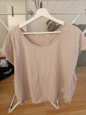 Ljusbeige t-shirt med blompatch - Enkel och snygg ljusbeige t-shirt med rund halsringning och korta ärmar. T-shirten har en liten ljusrosa blompatch nertill på framsidan. Perfekt till jeans eller shorts för en avslappnad look.