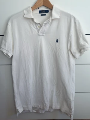 Vit pikétröja från Polo Ralph Lauren - Klassisk vit pikétröja från Polo Ralph Lauren med korta ärmar och blå broderad logga på bröstet. Tröjan har krage och knäppning vid halsen. Tillverkad i mjuk bomull som känns skön mot huden. Perfekt till jeans eller shorts. Säljer då för liten, mycket bra skick.