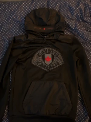 Svart hoodie från Zavetti Canada - Snygg svart hoodie från Zavetti Canada med stor huva och dragsko. På bröstet finns en sköld med en röd lönnlogga och mönstrad bakgrund. Praktisk magficka framtill och ribbade muddar vid ärmslut och nederkant. Perfekt för dig som gillar streetwear och vill ha något unikt. Pris kan diskuteras
