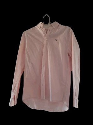 Ljusrosa skjorta från Gant - Klassisk ljusrosa skjorta från Gant med button-down krage och diskret logga broderad på bröstet. Skjortan har lång ärm, knappar framtill och är tillverkad i mjuk bomull. Perfekt för en clean och stilren look. Är igentligen i barn storlek men passar mig som är en M (skulle säga att den skulle sitta bäst på en S) Aldrig använd utan bara hängt i min garderob😅  (prövad 2 gånger)