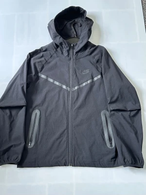 Svart vindjacka från Nike med huva - Svart vindjacka från Nike med hel dragkedja, två fickor med dragkedja och reflekterande detaljer över bröstet. Jackan har huva och elastiska muddar vid ärmsluten. Perfekt för blåsiga dagar och sportiga outfits.