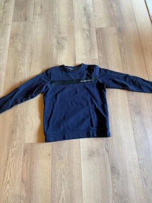 Mörkblå Sail Racing sweatshirt - Mörkblå sweatshirt från Sail Racing med svart rand över bröstet och logga tryckt på vänster sida. Tröjan har rund halsringning och långa ärmar. Perfekt för en avslappnad och sportig stil.