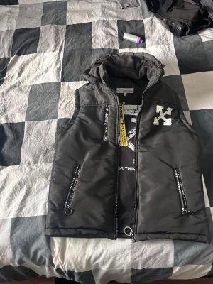 Svart dunväst från Off-White - Svart dunväst från Off-White med ikoniska vita pilar och texten 'SEEING THINGS' på ryggen. Västen har huva, dragkedjor med Off-White-tryck och en gul tag. Perfekt för dig som gillar streetwear och vill sticka ut med en snygg och modern look.