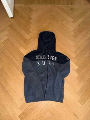 Mörkblå Hollister hoodie med dragkedja - Snygg mörkblå hoodie från Hollister med dragkedja framtill och stor huva. Framsidan har trycket 'HOLLISTER SURF' i vitt och på ärmen sitter en brun läderpatch med Hollister-loggan. Tillverkad i mjukt bomullsmaterial, perfekt för chill dagar.