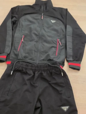 Svart och vit tracksuit set från Prada - Säljer två tracksuit set från Prada, ett i svart med grå detaljer och röda dragkedjor, och ett i vitt med svarta detaljer. Båda seten har jacka med dragkedja, sidofickor och matchande byxor med resår i midjan. Diskret Prada-logga på både jacka och byxa.