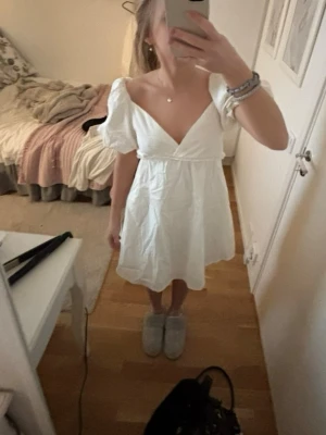 Vit kort klänning/studentklänning - Söt vit kortklänning med puffärmar och djup v-ringning. Klänningen har en omlott-look upptill och markerad midja som ger en feminin siluett. Perfekt för sommarens alla tillfällen. Materialet känns lätt och luftigt. Från Nelly aldrig använd