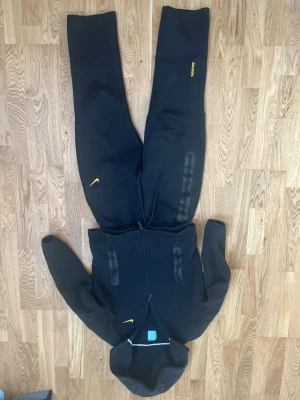 Svart Nike jumpsuit med huva - Säljer en svart jumpsuit från Nike med huva och dragkedja framtill. Jumpsuiten har gula detaljer som Nike-loggan och text på benet. Tillverkad i mjukt och bekvämt material, perfekt för chill dagar. Justerbar midja med snörning och långa ärmar.