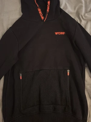 Svart hoodie från WOXO 720° - Svart hoodie från WOXO 720° med orange logga på bröstet och detaljer på huvan. Stor ficka framtill med mesh och dragkedjor. Skön passform och sportig look, perfekt för dig som gillar streetwear och vill sticka ut lite extra.