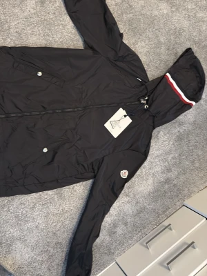 Svart vindjacka från Moncler - Svart vindjacka från Moncler med huva och dragkedja framtill. Jackan har två fickor med tryckknappar och en vit-röd rand på huvan. Moncler-logga på ena ärmen. Perfekt för blåsiga dagar och snygg streetstil.