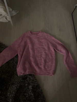 Rosa stickad tröja med rund hals - Mysig rosa stickad tröja med rund hals och långa ärmar. Tröjan har en enkel design och är tillverkad i ett mjukt material som passar perfekt för kyligare dagar. Ribbstickade muddar vid ärmslut och nederkant ger en snygg finish.